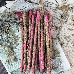 Puede incluir: Un manojo de varitas de incienso largas, finas y de color rosa brillante, cubiertas de hierbas secas y pétalos de flores. Las varitas están dispuestas verticalmente sobre una colección de páginas de libros botánicos antiguos con texto e ilustraciones.