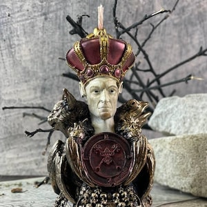 Puede incluir: Una detallada escultura de vela que representa una figura coronada con una expresión seria. La figura está flanqueada por un gato y una rana, sosteniendo un escudo con un símbolo. La vela tiene una combinación de colores burdeos y dorado, con una mecha en la parte superior.