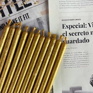 Puede incluir: Un paquete de 15 velas de té doradas. Las velas están dispuestas en fila y todas son del mismo tamaño. Las velas están sobre un fondo de periódico.