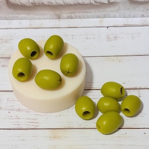 May include: A selection of green olive-shaped items, some placed on a white, circular base. The olives are a vivid green and have a hole at one end. The background includes a white brick wall and a wooden surface.
