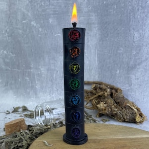 May include: Black candle with seven colorful chakra symbols, lit and burning. The candle is on a wooden surface with dried herbs and a glass bottle.
