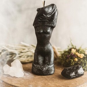 Black handcrafted candle shaped like a stylized female body torso, displayed on a wooden stand with crystals and dried herbs in a mystical setup.