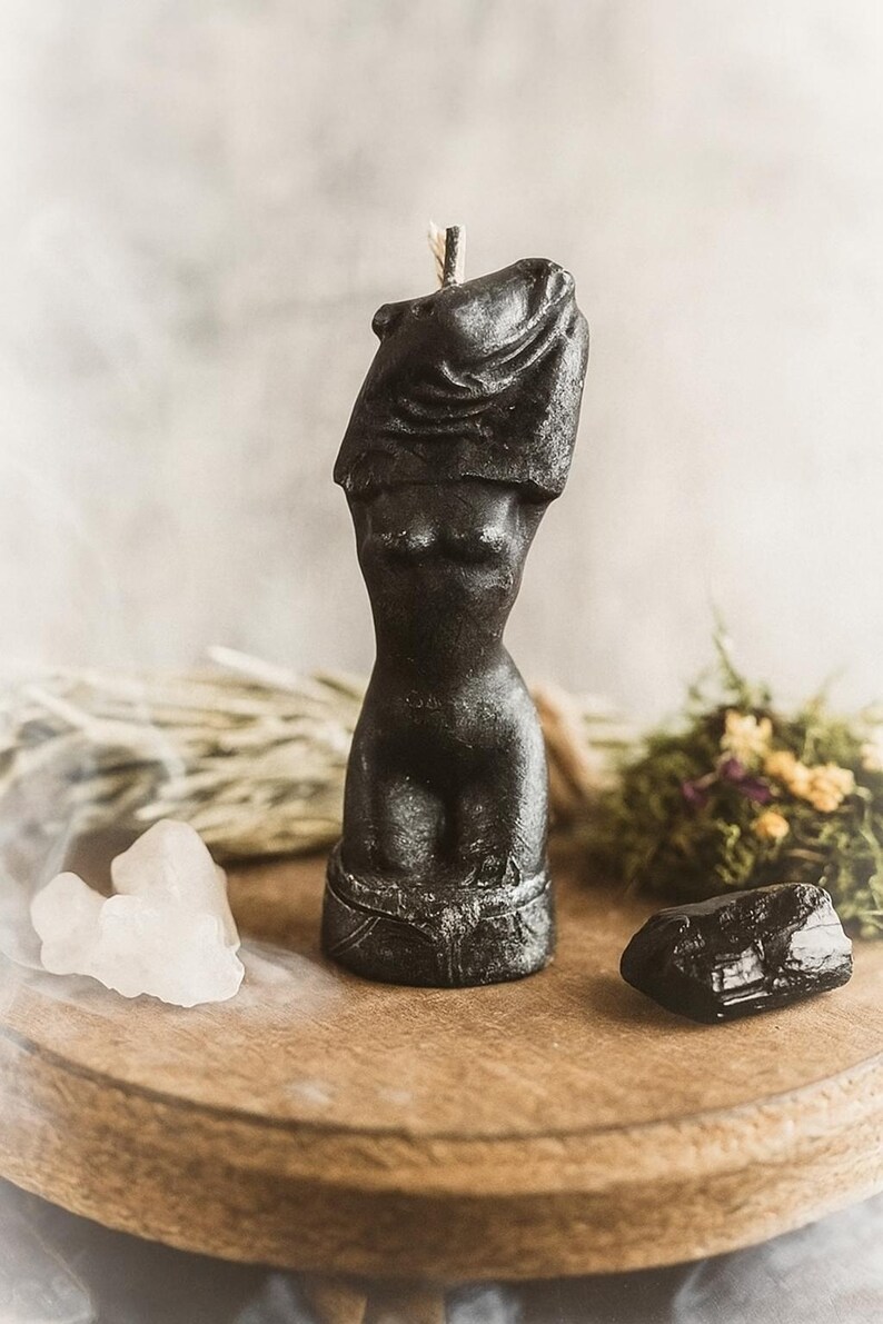 Black handcrafted candle shaped like a stylized female body torso, displayed on a wooden stand with crystals and dried herbs in a mystical setup.