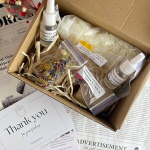 Open kraft box with soy wax flakes, dried botanicals, fragrance bottles, and dye packets arranged neatly inside with shredded filler, styled beside a "Thank you" card and red flowers.