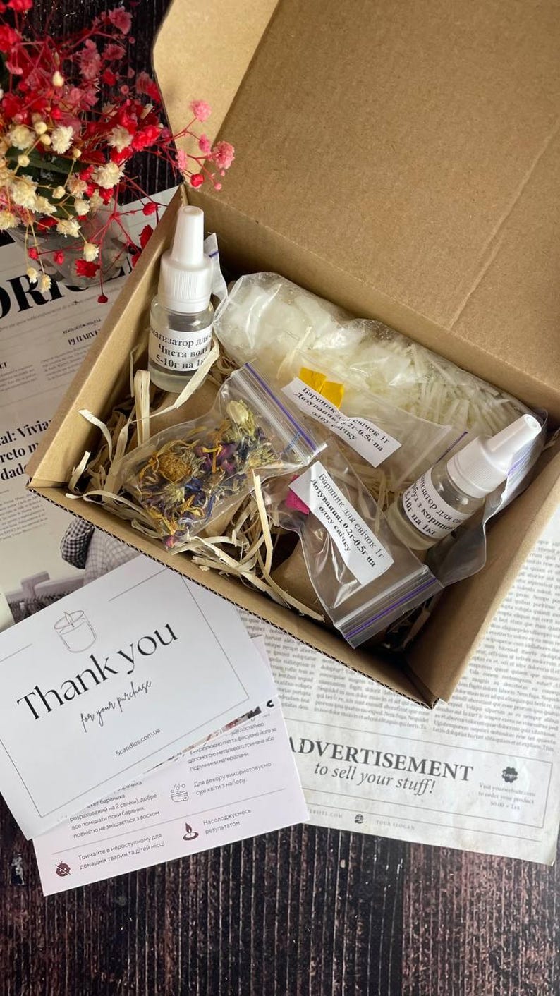 Open kraft box with soy wax flakes, dried botanicals, fragrance bottles, and dye packets arranged neatly inside with shredded filler, styled beside a "Thank you" card and red flowers.
