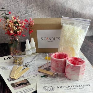 Full soy candle making kit displayed on table: red marble jars, soy wax bag, fragrance oils, wooden wicks, dyes, dried flowers, and tools beside branded box and instruction sheets.
