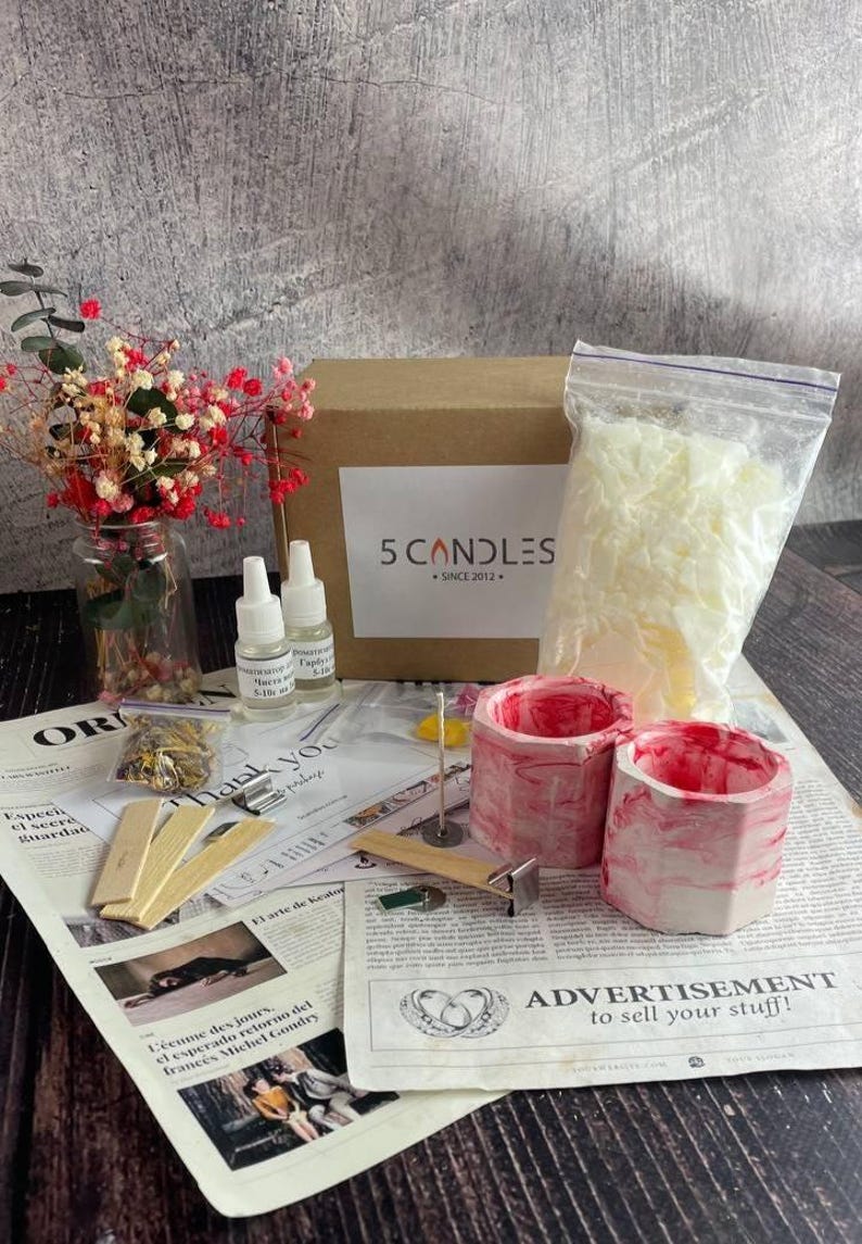 Full soy candle making kit displayed on table: red marble jars, soy wax bag, fragrance oils, wooden wicks, dyes, dried flowers, and tools beside branded box and instruction sheets.