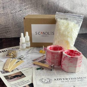 Full soy candle making kit displayed on table: red marble jars, soy wax bag, fragrance oils, wooden wicks, dyes, dried flowers, and tools beside branded box and instruction sheets.