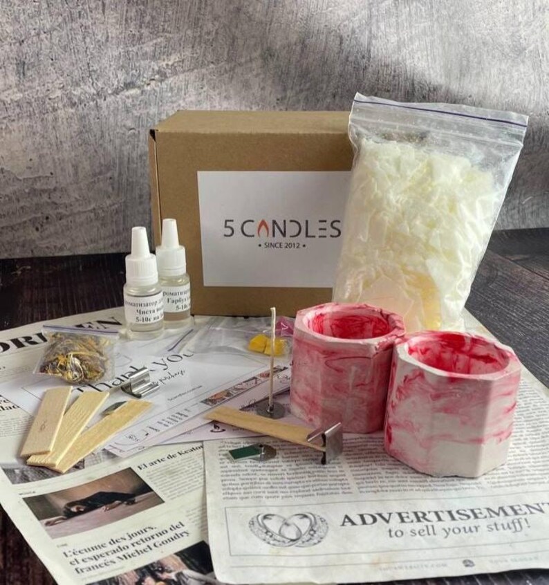 Full soy candle making kit displayed on table: red marble jars, soy wax bag, fragrance oils, wooden wicks, dyes, dried flowers, and tools beside branded box and instruction sheets.