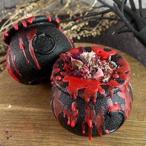 Könnte beinhalten: Zwei schwarze, kesselartige Kerzen mit rotem Wachs, das an den Seiten heruntertropft. Ein Kessel enthält getrocknete rote Blumen und Kräuter.