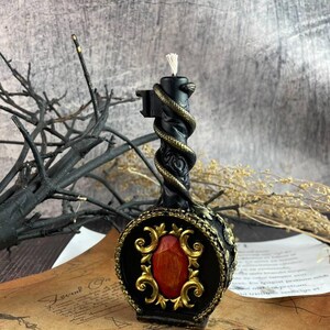 Black serpent-wrapped candle with gold detailing and a red gem centerpiece, placed on a tarot and parchment setup with red stones and mystical altar elements.