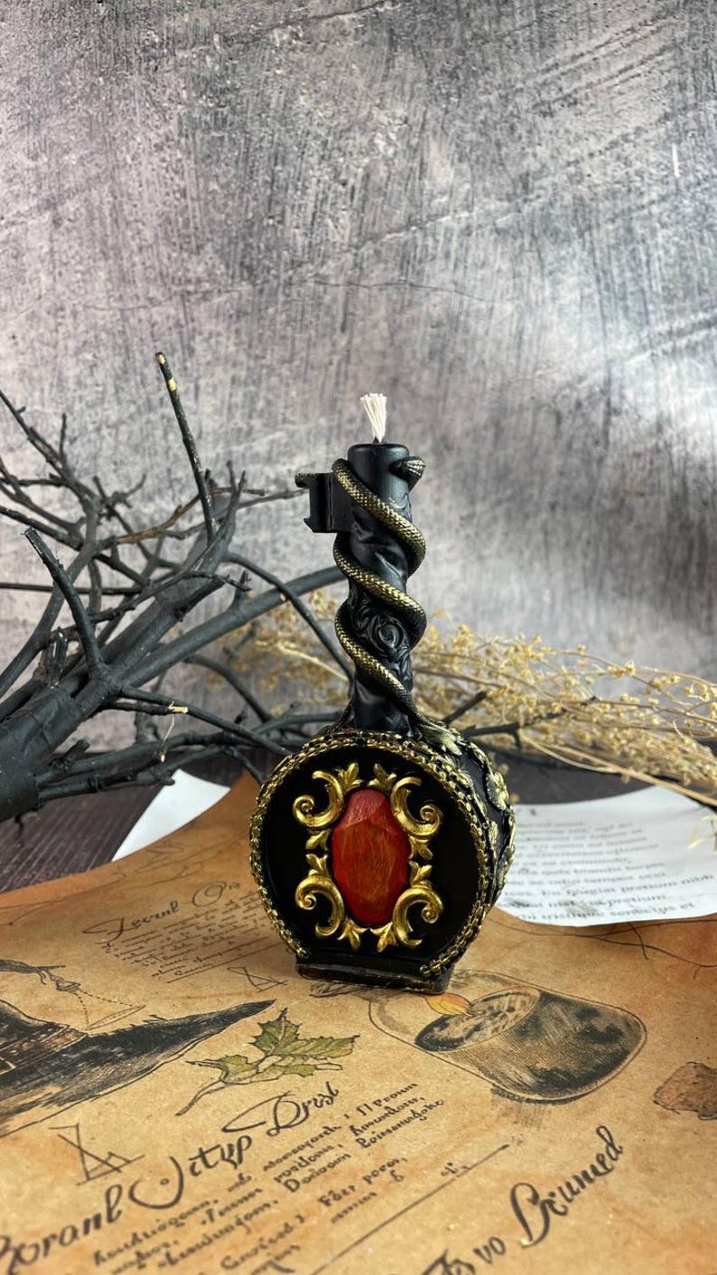 Black serpent-wrapped candle with gold detailing and a red gem centerpiece, placed on a tarot and parchment setup with red stones and mystical altar elements.