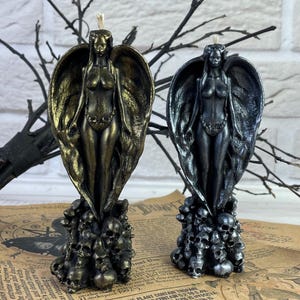 Puede incluir: Dos velas de ángel negras y doradas con alas y una figura femenina de pie sobre una base de cráneos. Las velas están sobre una superficie de papel impreso.