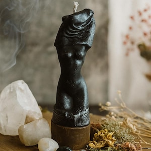 Black wax candle shaped as a female torso standing on a wooden altar surrounded by crystals, herbs, and smoke, symbolizing feminine power and beauty.