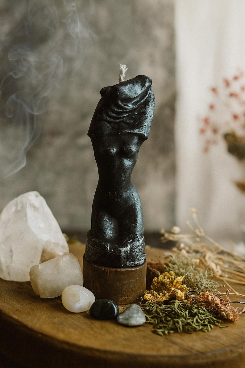 Black wax candle shaped as a female torso standing on a wooden altar surrounded by crystals, herbs, and smoke, symbolizing feminine power and beauty.