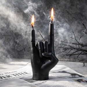 Black sculptural candle shaped like a rock hand gesture with two flames burning, surrounded by smoke and branches, symbolizing rebellion, strength, freedom, and raw inner power.