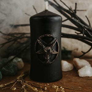 Black ritual candle with a bronze Baphomet emblem, placed on a wooden altar surrounded by white crystals, dried herbs, and dark branches.