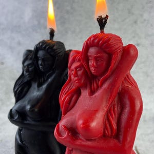 Könnte beinhalten: Zwei geformte Kerzen, eine rote und eine schwarze, zeigen Figuren, die sich umarmen. Beide Kerzen sind mit Flammen angezündet.