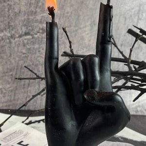 Black wax candle in the shape of a hand showing the rock horns gesture with flames on both fingertips, minimal background
