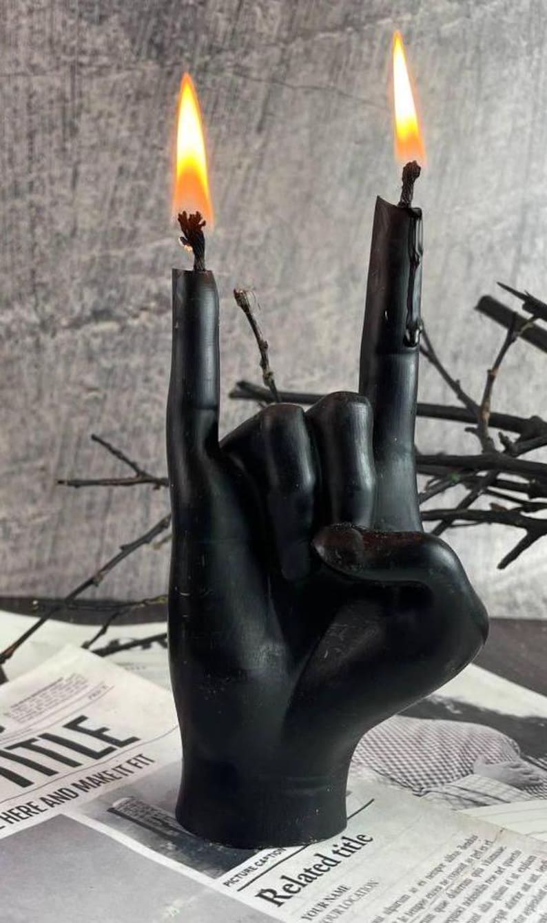 Black wax candle in the shape of a hand showing the rock horns gesture with flames on both fingertips, minimal background