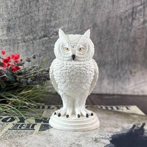 May include: A white owl figurine with detailed feather textures and yellow eyes. The owl is perched on a round base with black accents on its feet. The background features a textured gray surface and floral accents, suggesting a decorative item.