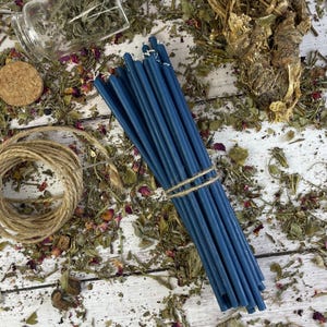 Puede incluir: Un paquete de velas de color azul oscuro atadas con cuerda. Las velas están sobre una superficie blanca con hierbas y flores secas esparcidas a su alrededor.