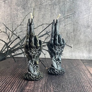 May include: Two black hand-shaped candles with intricate white designs and the word "Solstice" on the base. The candles are lit and have a gothic aesthetic.