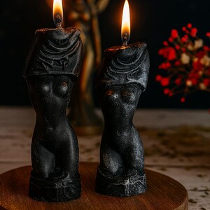 Two black sculptural candles shaped like female torsos with veiled heads, lit and standing on a wooden round platform, with a goddess statue and dried flowers in the background.