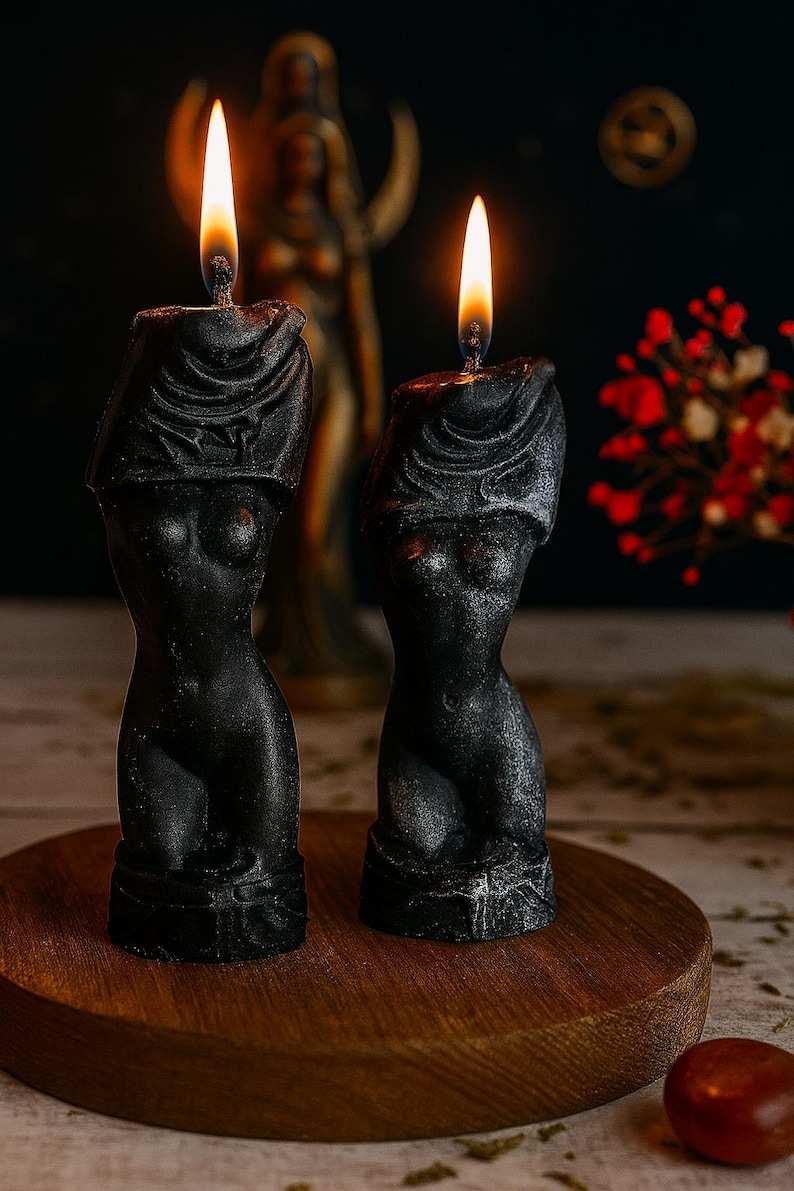 Two black sculptural candles shaped like female torsos with veiled heads, lit and standing on a wooden round platform, with a goddess statue and dried flowers in the background.