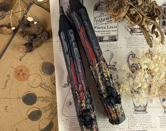 May include: Two black and red candles with gold accents. The candles are long and thin, and they are sitting on a piece of paper with text that says "READ THE PREFACE BEFORE ORDERING."