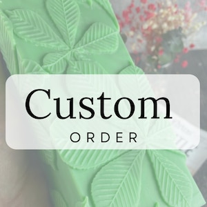 Può includere: Un oggetto rettangolare verde chiaro con motivi a forma di foglia e le parole "Custom ORDER" in una scatola bianca. L'oggetto ha una superficie strutturata con disegni di foglie in rilievo. Lo sfondo è sfocato, con accenni di fiori.