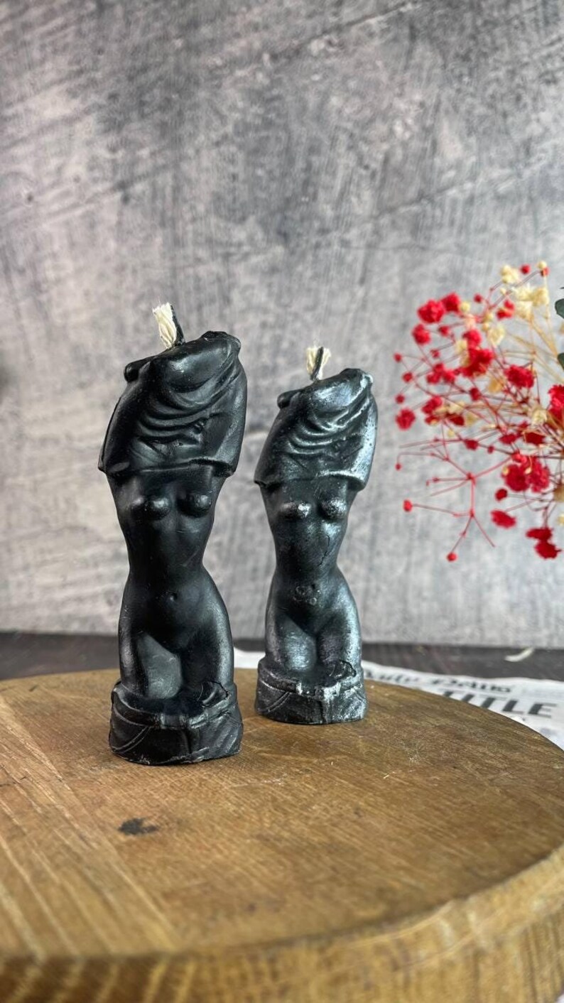 Two black handcrafted female body candles styled like classical sculptures, placed on a round wooden platform next to a glass vase with red and white roses