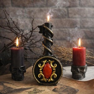 Black ritual candle shaped like a mystical key with ornate red gemstone detail, surrounded by burning candles on skull holders and smoky atmospheric altar setting.
