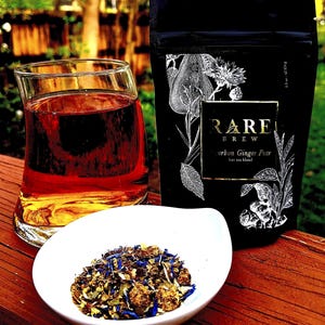 May include: A glass of tea with a white ceramic bowl filled with loose leaf tea. The tea bag packaging is black with gold lettering and says "RARE BREW urban Ginger Pear hot tea blend".