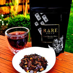 May include: A black pouch with gold lettering that reads "RARE BREW" and "Dark Chocolate Cherry loose tea blend". The pouch is sitting on a wooden surface next to a glass cup of tea and a white bowl filled with loose leaf tea.