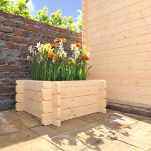 May include: A wooden planter box with a light brown finish, filled with a variety of yellow, orange, and white flowers. The planter is sitting on a patio with a brick wall in the background.