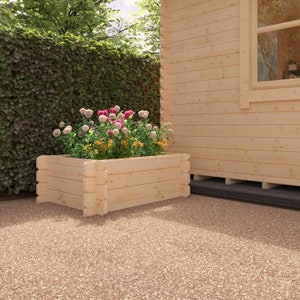 May include: A wooden planter box, filled with pink, white, and yellow flowers, sits on a speckled surface. The light-coloured wooden planter is near a wooden building and a green hedge, creating a garden scene.