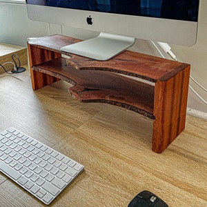 May include: A wooden monitor stand with two shelves, made from natural wood with a rich brown color and a unique, curved design. The stand is perfect for elevating a computer monitor and creating a more ergonomic workspace.
