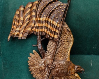 May include: A wooden carving of an eagle holding an American flag. The eagle is brown and gold, and the flag is red, white, and blue. The carving is on a green background.