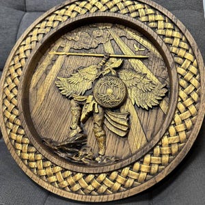May include: A round, wooden wall art piece with a detailed relief carving. The artwork features a warrior figure with wings, holding a spear and shield. The piece has a woven border and is finished in shades of gold and brown.