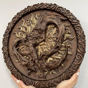 Op de afbeelding: Een ronde houten wandplaquette met een gedetailleerde carving van een vrouw en een draak in een vurige omhelzing. De plaquette is versierd met ingewikkelde krulwerk rond de rand en heeft een gouden afwerking.