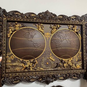 May include: A dark brown, ornate framed wall art piece featuring two detailed world maps. The maps are encircled with gold accents and surrounded by gold relief figures. The frame has intricate carvings.