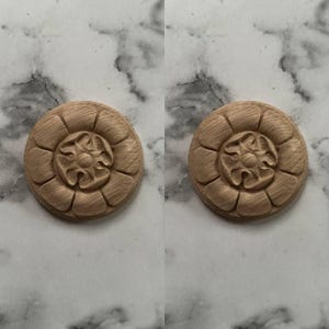 May include: Two wooden, circular decorative appliques. Each applique features a central floral design surrounded by a series of curved, petal-like shapes. The wood has a natural, light brown color, and the details are carved.