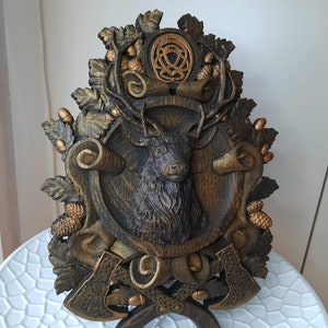 May include: A wooden wall plaque with a carved deer head, surrounded by leaves and acorns. Two crossed axes are at the bottom of the plaque, with a Celtic knot design at the top.