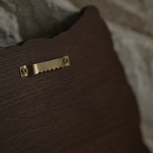 Puede incluir: Una placa de madera marr&oacute;n oscuro con un gancho de metal dorado en la parte posterior.
