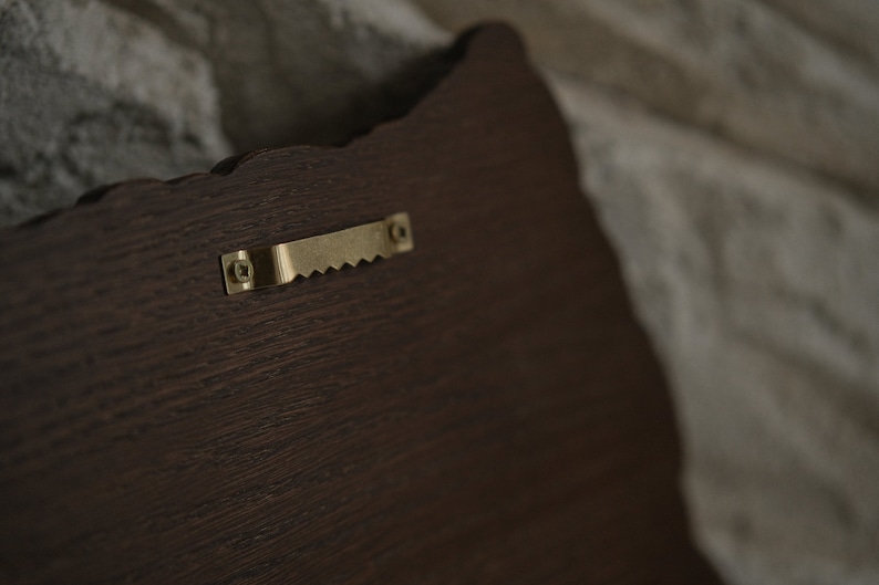 Puede incluir: Una placa de madera marr&oacute;n oscuro con un gancho de metal dorado en la parte posterior.