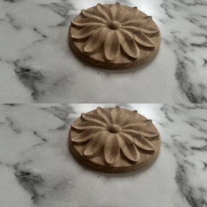May include: Two wooden, circular decorative elements with a carved floral design. The elements are light brown and appear to be made of wood. The background is a marble-like surface.