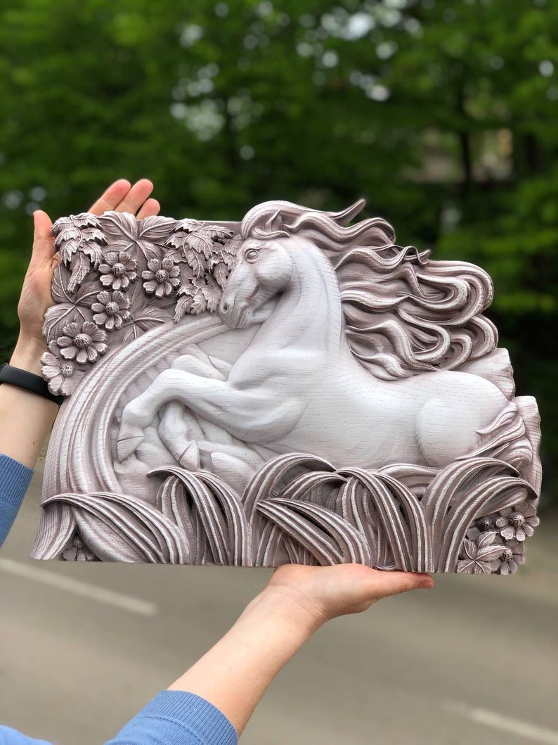 May include: A white wooden wall sculpture of a horse running through a field of flowers and grass. The horse has a flowing mane and tail. The sculpture is painted in a light brown colour.