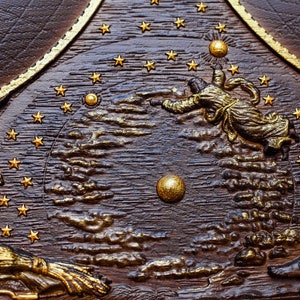 Puede incluir: Una placa de madera marr&oacute;n oscuro con un dise&ntilde;o pintado en oro que representa una escena celestial. La placa presenta un c&iacute;rculo central con un sol y estrellas pintados en oro, rodeados de nubes y figuras. Las figuras est&aacute;n representadas de forma estilizada, con una figura que alcanza el sol.