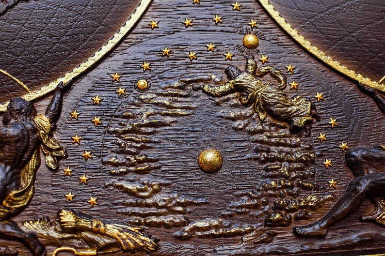 Puede incluir: Una placa de madera marr&oacute;n oscuro con un dise&ntilde;o pintado en oro que representa una escena celestial. La placa presenta un c&iacute;rculo central con un sol y estrellas pintados en oro, rodeados de nubes y figuras. Las figuras est&aacute;n representadas de forma estilizada, con una figura que alcanza el sol.